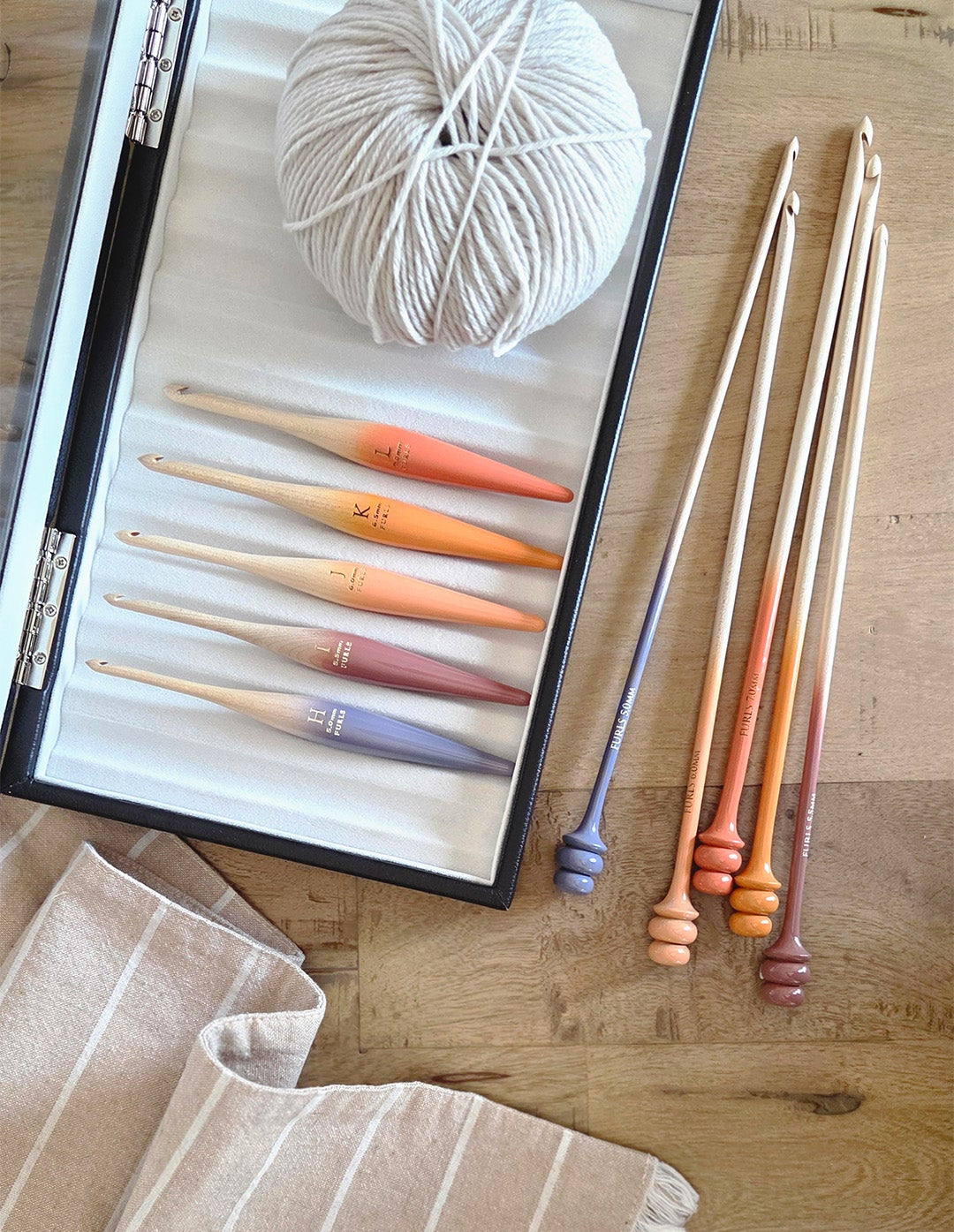 A knitting or crocheting kit laid out on a wooden surface, including a ball of white yarn, several wooden crochet hooks with colorful tips, and a folded beige striped fabric.